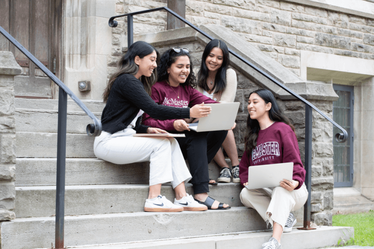 Students sitting on University Hall steps pointing to a laptop screen.